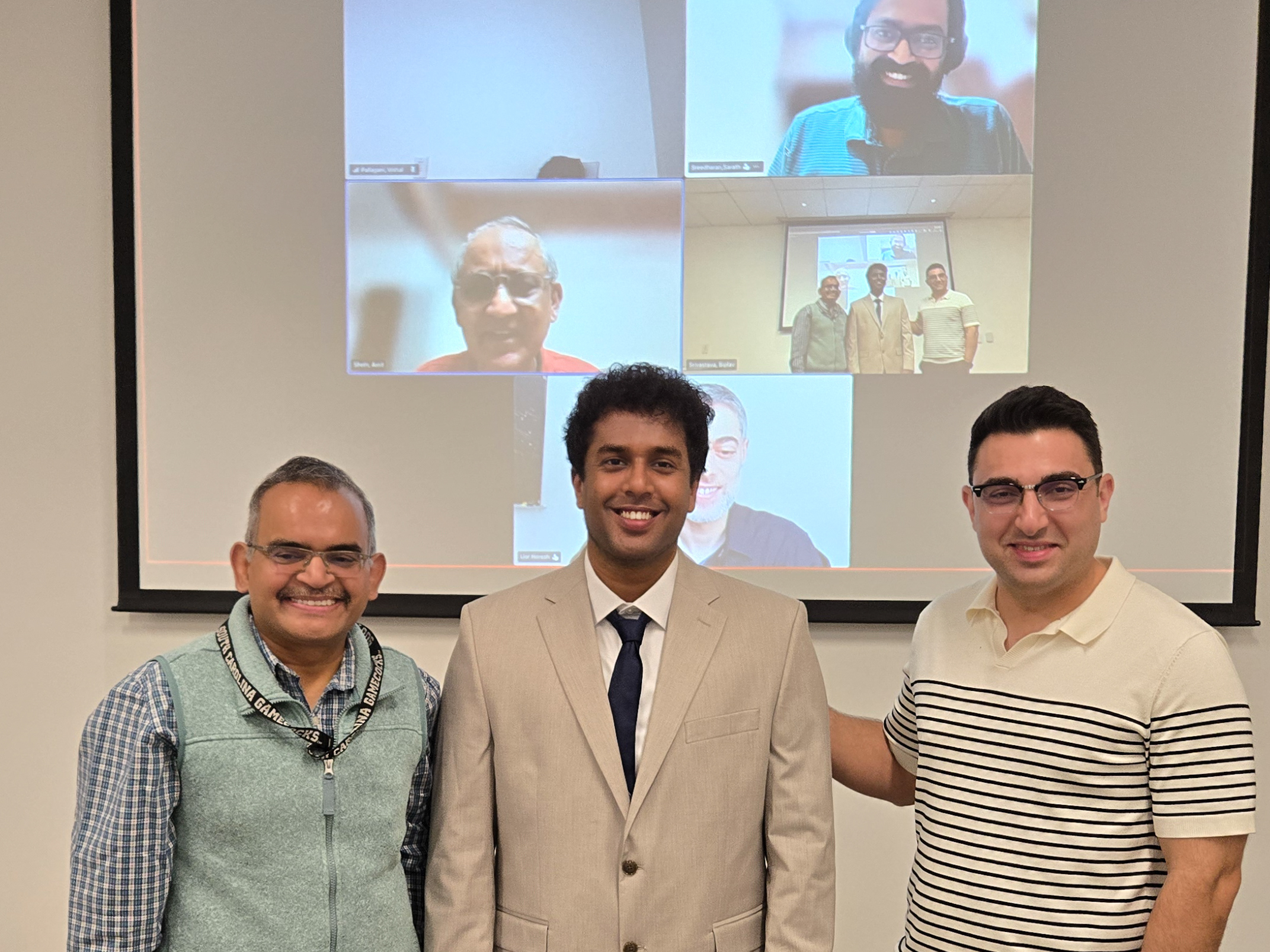 Vishal Pallagani posing after the defense with attendees in the room and remote participants on screen.